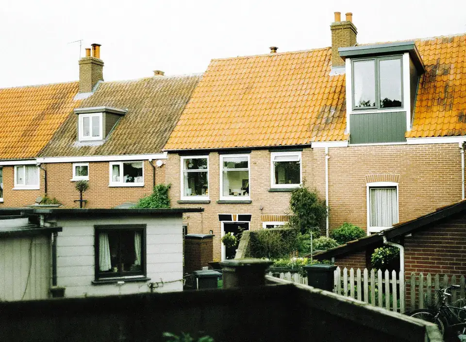 De achterkant van een rij bakstenen jaren-30-woningen met oranje pannendaken, schoorstenen, een dakkapel en kleine tuinen met schuttingen