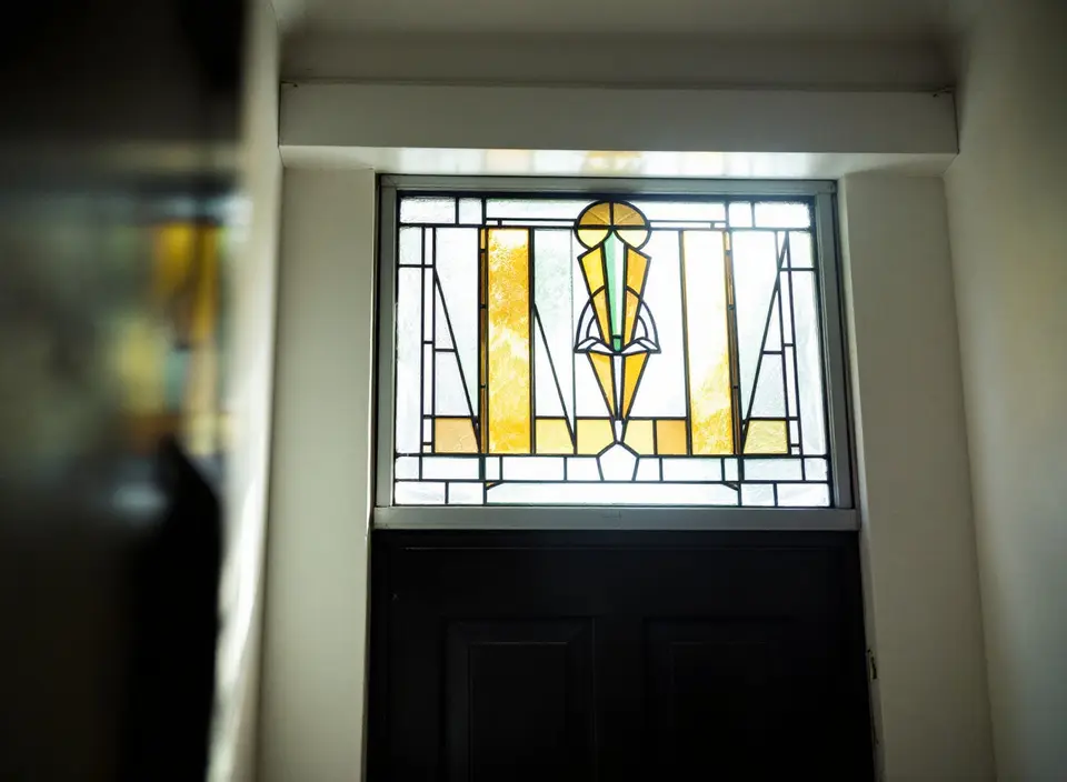 Een bovenlicht van glas-in-lood boven een voordeur, van binnen gezien, met een geometrisch patroon in amber, groen en helder glas