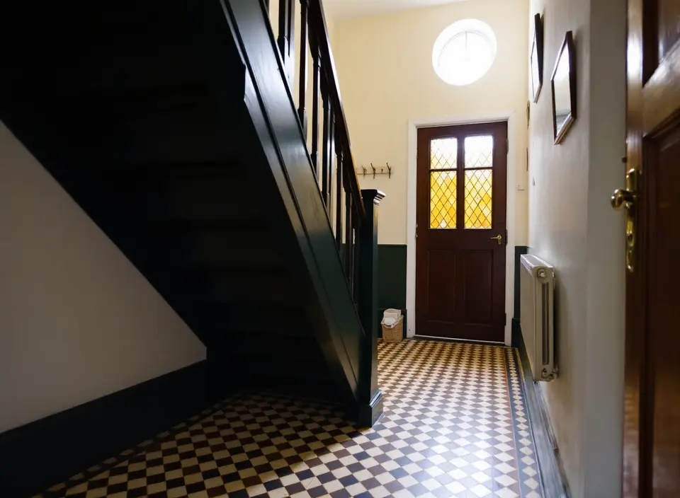 De smalle gang van een jaren-30-woning met een vloer van kleine geometrische tegels, een donkere houten trap en een voordeur met geel glas-in-lood